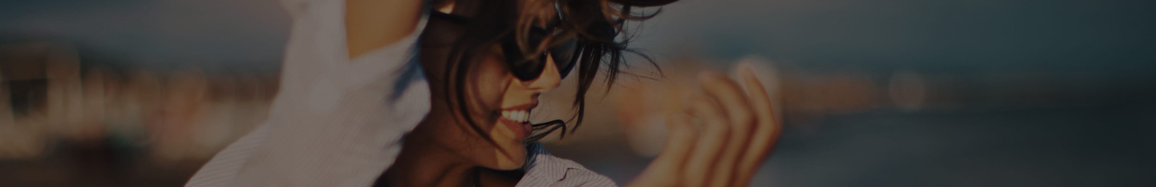 A person with long hair smiles and raises their hand against a blurred outdoor background