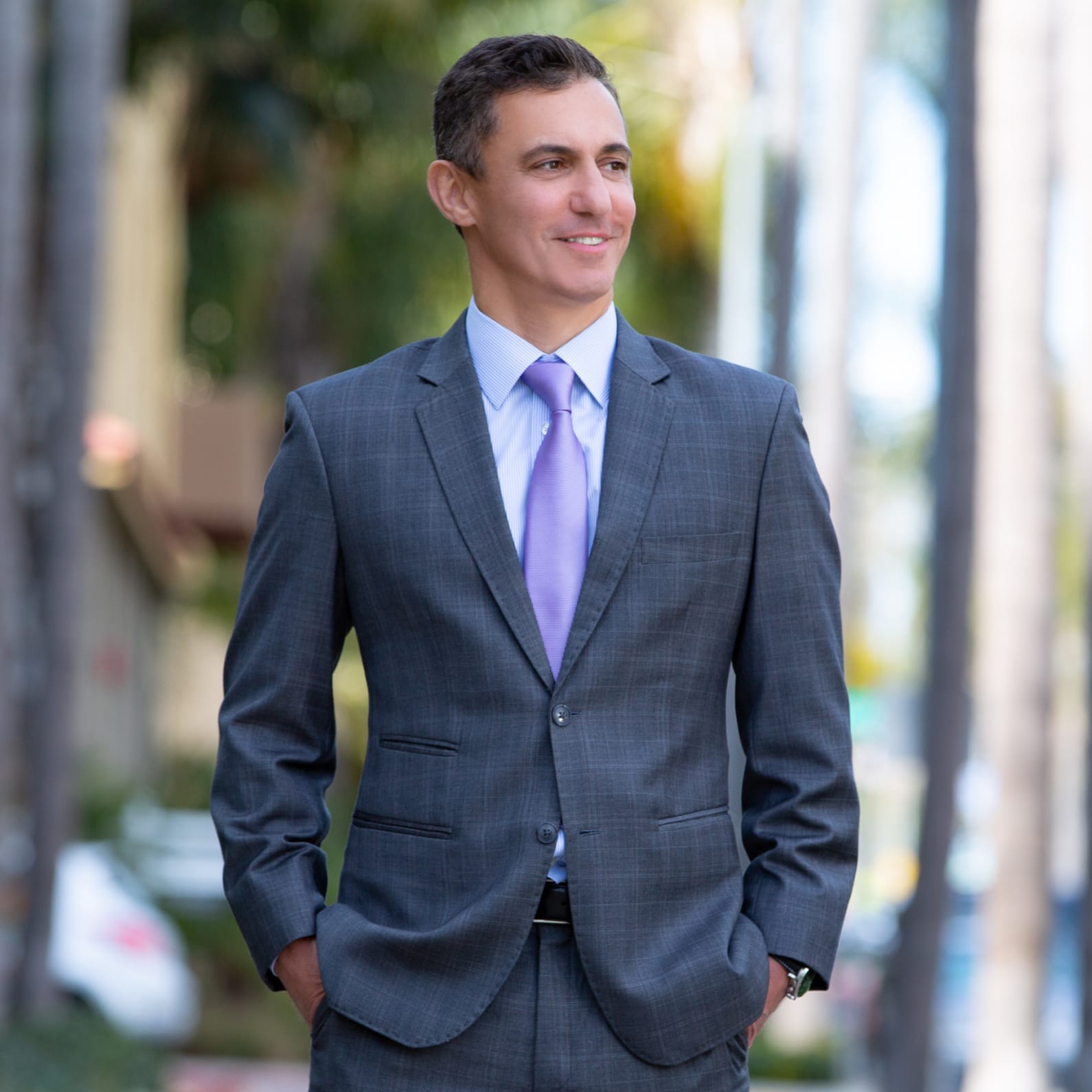 Man in a gray suit and purple tie, standing outdoors with hands in pockets