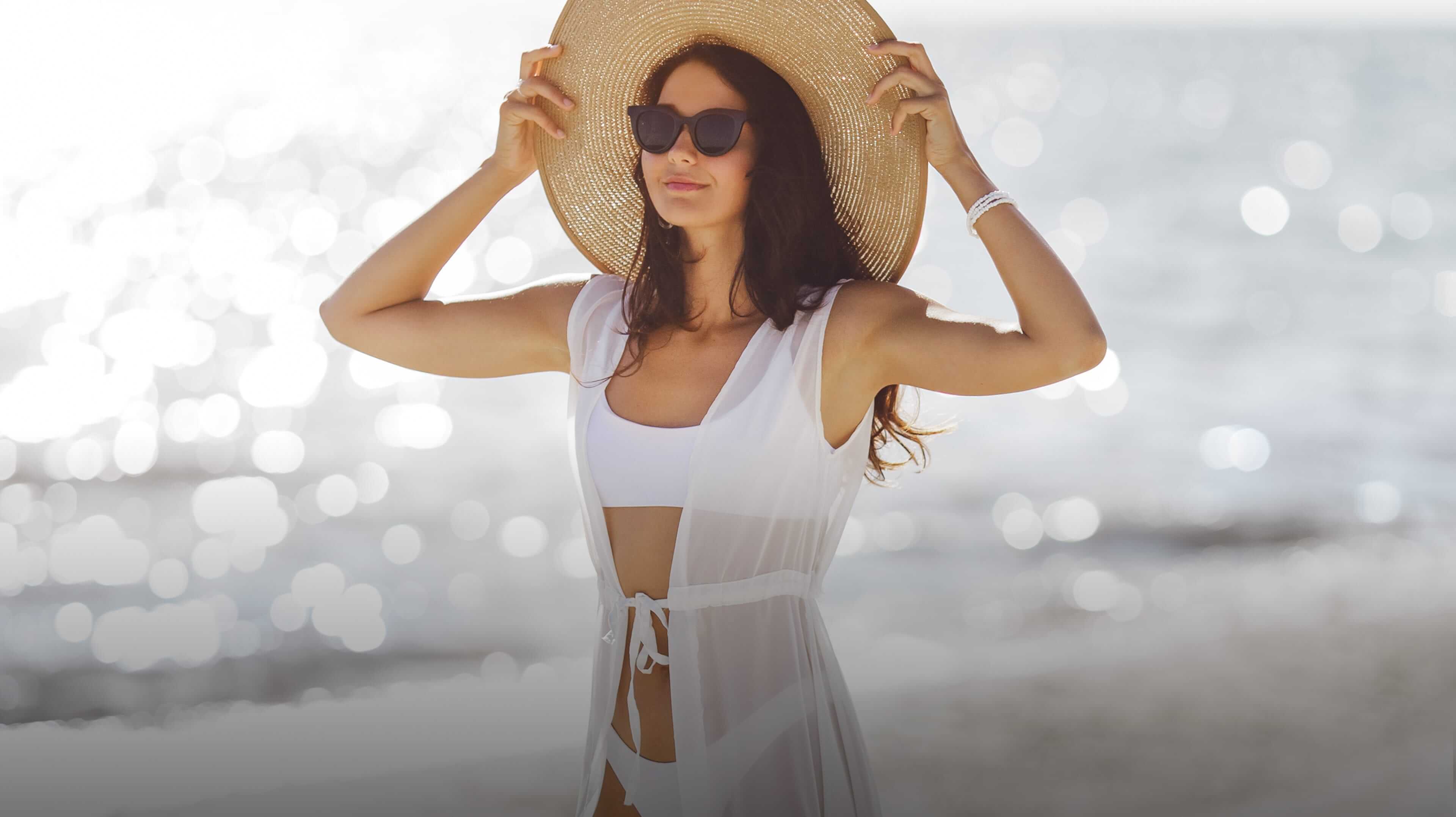 A woman in a white swimsuit and hat stands on the beach, sunglasses on, looking around