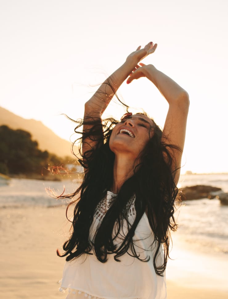 A woman with long hair smiles and raises her arms at the beach during sunset