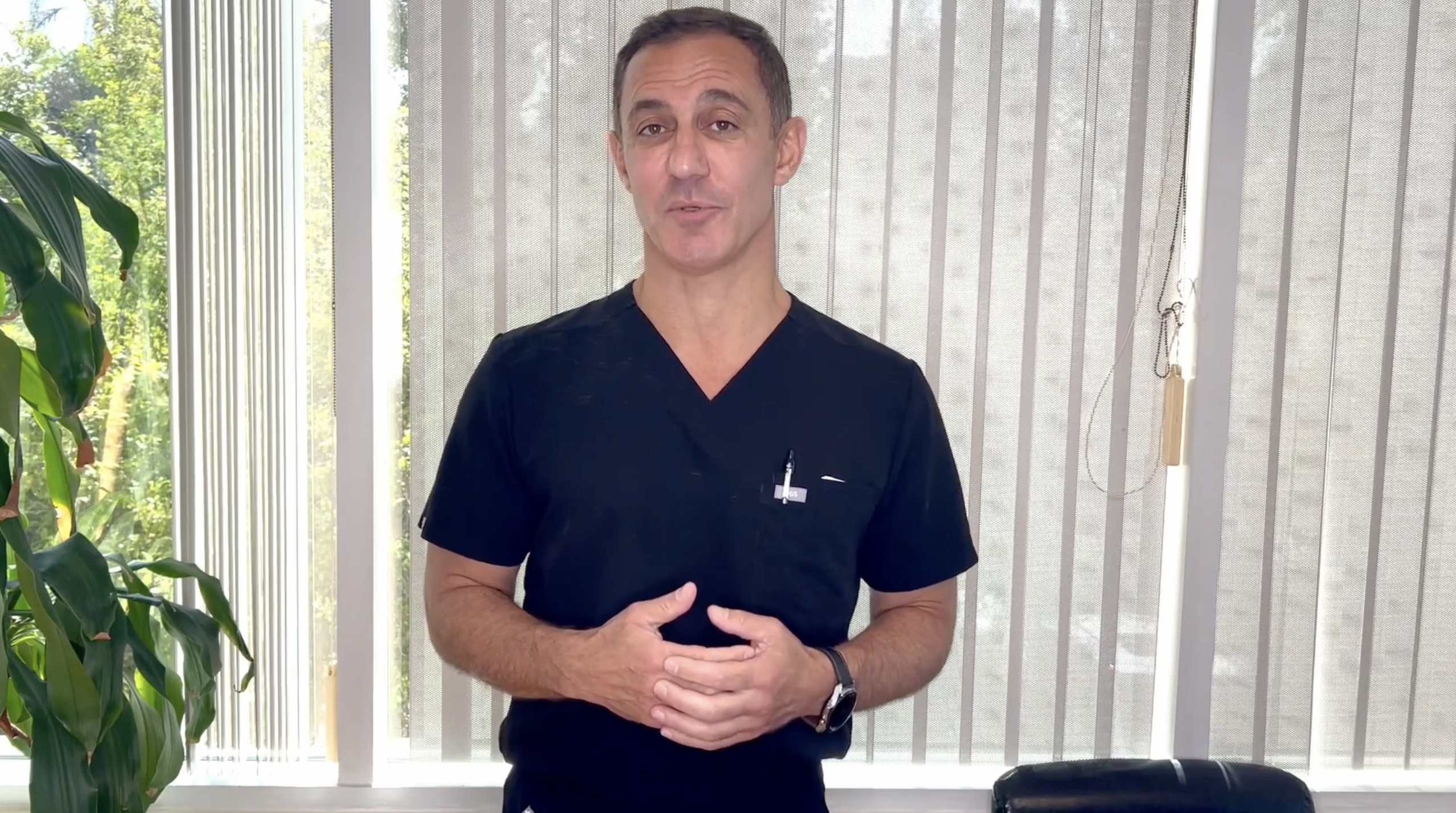 A man in scrubs stands in an office with large windows and plants