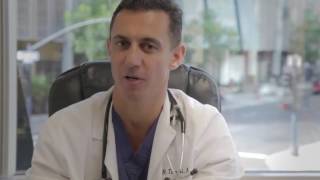 A doctor sitting in a chair, wearing a white coat and stethoscope, speaking to the camera