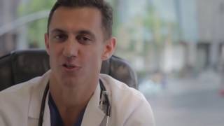 A man wearing a white coat and stethoscope sits in an office, speaking