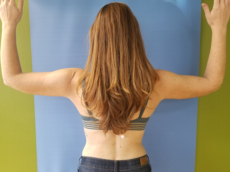 A person with long hair stands facing away with arms raised against a blue background