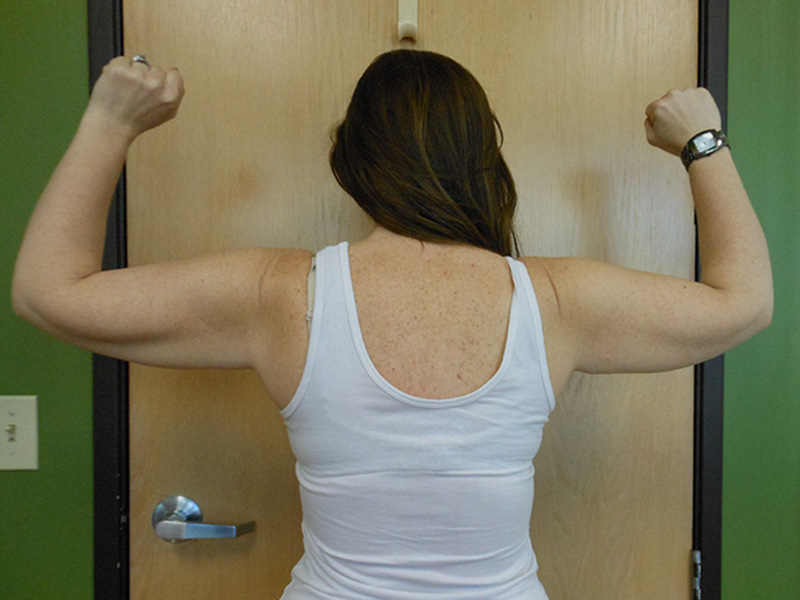 A woman stands with her back to the camera, flexing her arms. A door is in the background