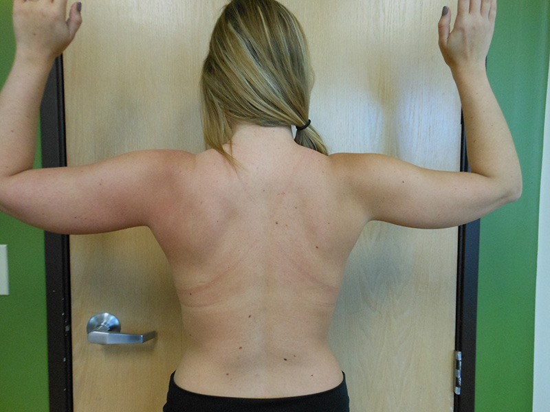 Person with long hair facing a wall, arms raised, back visible, standing indoors