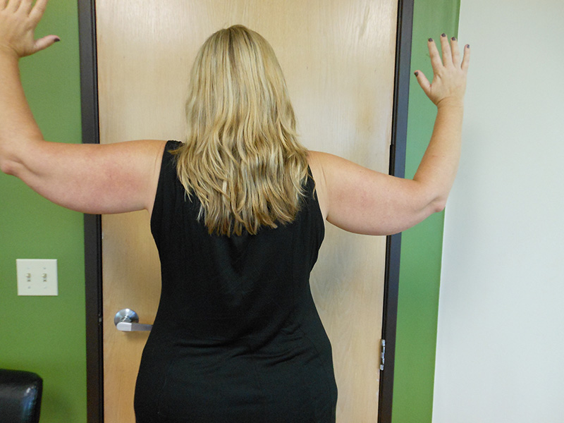 A person with long hair stands facing a closed door, hands raised against the wall