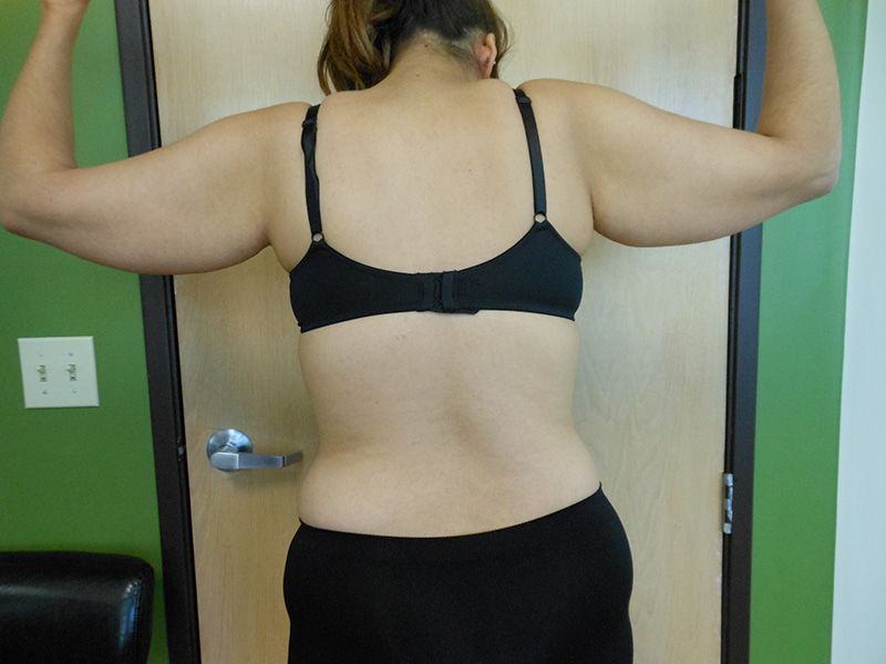 A person with shoulder-length hair stands with arms raised, facing a wall