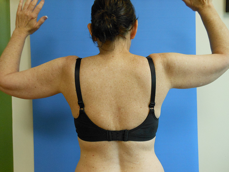A woman from the back raises her arms, wearing a black bra against a blue background