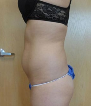 A side view of a person in a black bra and blue underwear standing against a wooden wall